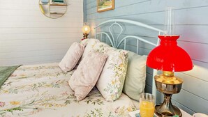 Un dormitorio con una cama, ropa de cama floral, una lámpara roja, una mesita con un vaso de jugo de naranja y un estante en la pared con libros y un florero.
