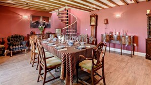 Uma sala de jantar com uma mesa comprida posta para seis pessoas, cadeiras de madeira, uma escada em espiral e um relógio grande na parede.