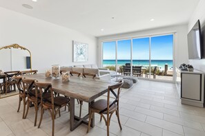 Ein geräumiger Essbereich mit einem großen Holztisch und Stühlen, einem weißen Sofa und Blick auf den Strand durch große Fenster.