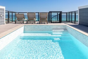 Ein Pool auf dem Dach mit klarem, blauem Wasser, drei Liegestühlen und Blick auf den Ozean.