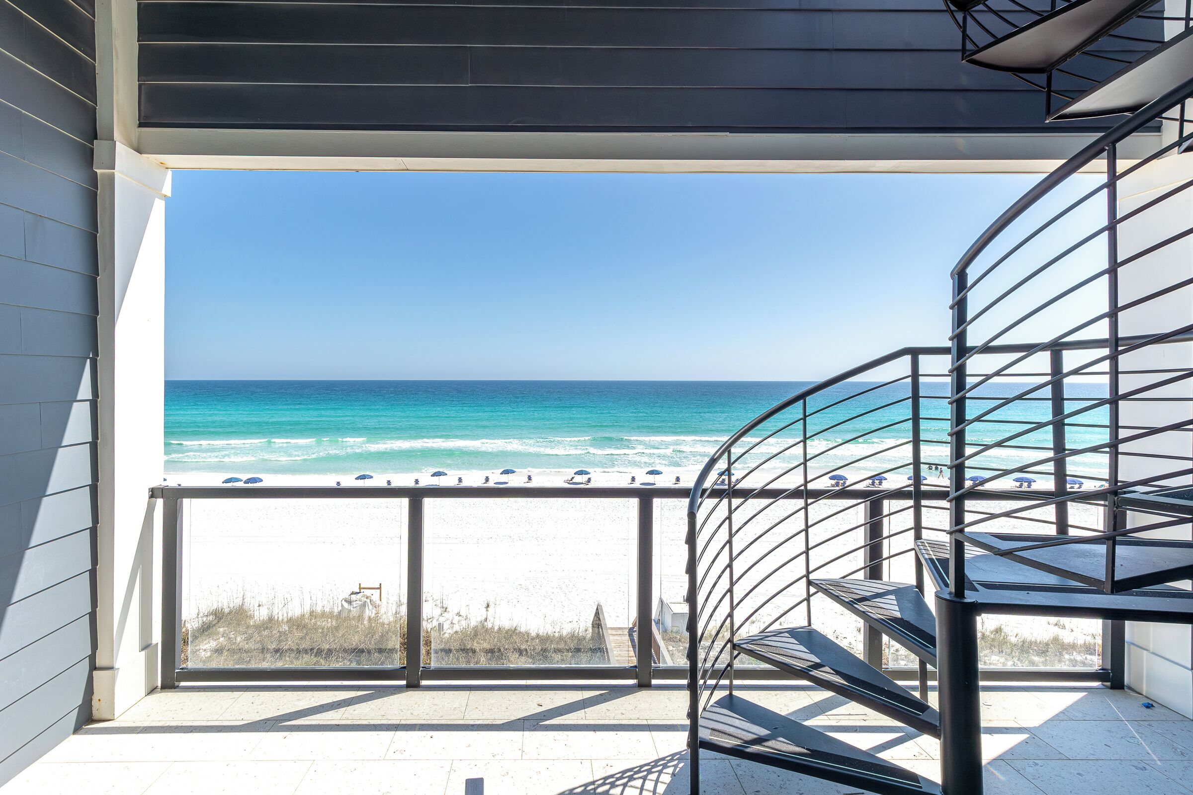 A balcony with a view of the ocean and beach, a metal spiral staircase, and a clear sky.
