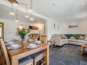 A dining area with a wooden table set for four, surrounded by chairs, and a living area with a sofa and a coffee table. - 4 Bed in Bude (oc-91927) (Hobbacott)