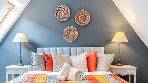 Ein Schlafzimmer mit einem grau gepolsterten Kopfteil, einer farbenfrohen, gestreiften Decke und zwei Nachttischlampen.