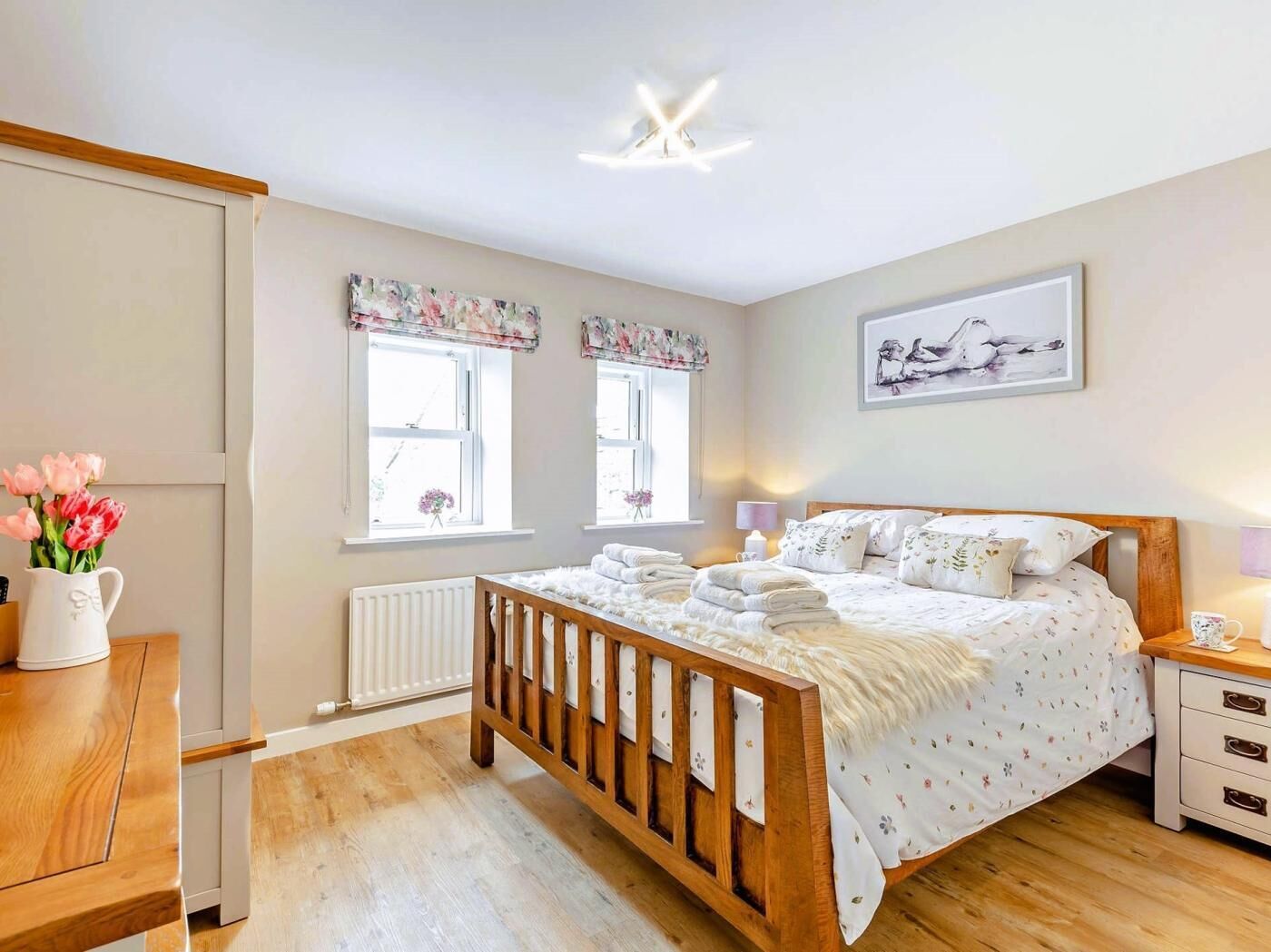 A bedroom with a wooden bed, two windows with floral curtains, a radiator, a bedside table with a lamp, and a framed picture on the wall.