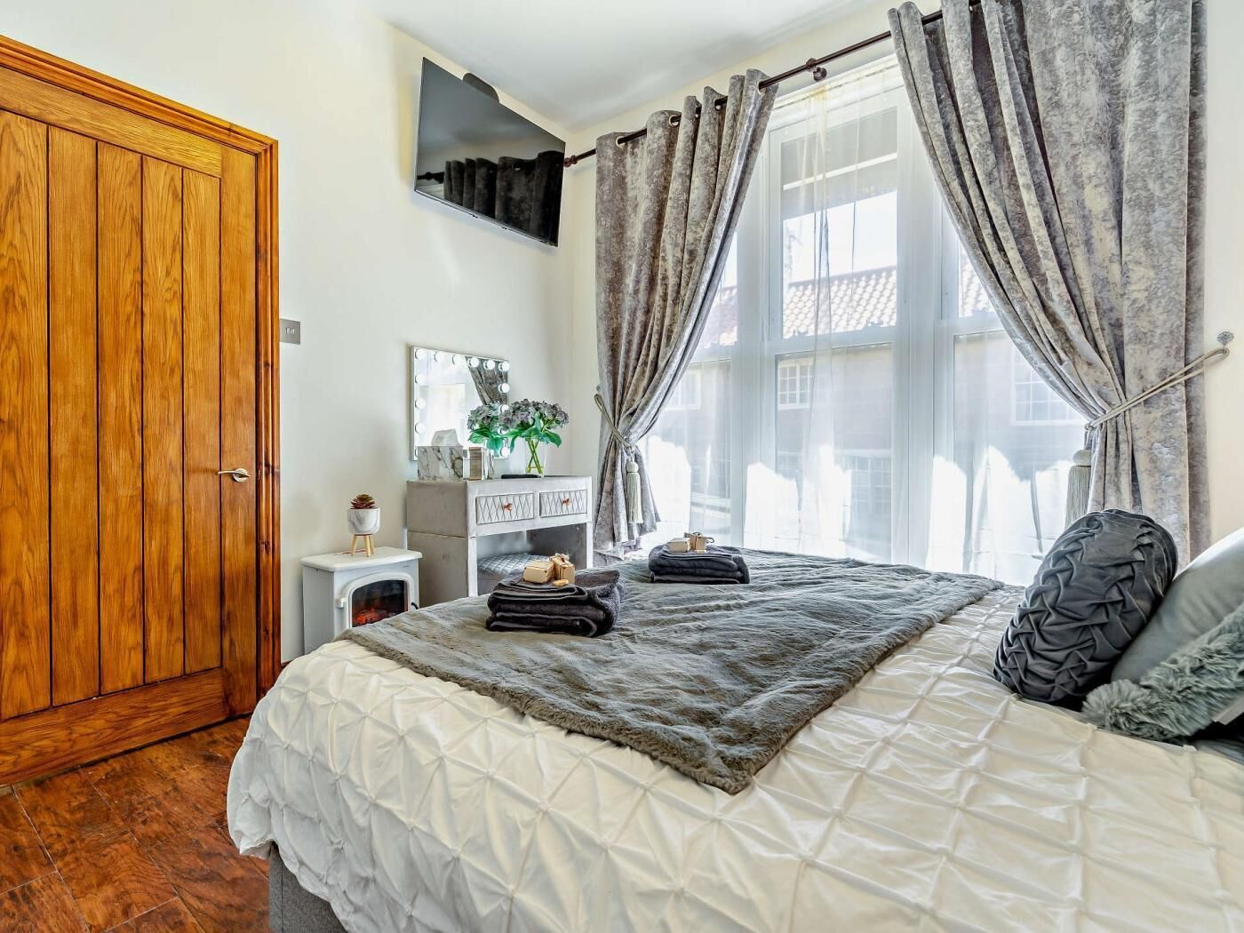 A bedroom with a wooden wardrobe, a vanity with a mirror, a bedside table, a bed with a grey blanket, and a window with curtains.