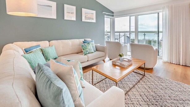 Een moderne woonkamer met een bank, fauteuils, een salontafel en uitzicht op de zee door grote ramen.