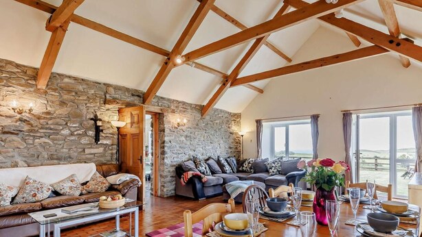 Ein gemütliches Wohnzimmer mit Steinwand, Holzbalken, einem Sofa, einem Esstisch für vier Personen und Blick durch ein großes Fenster.