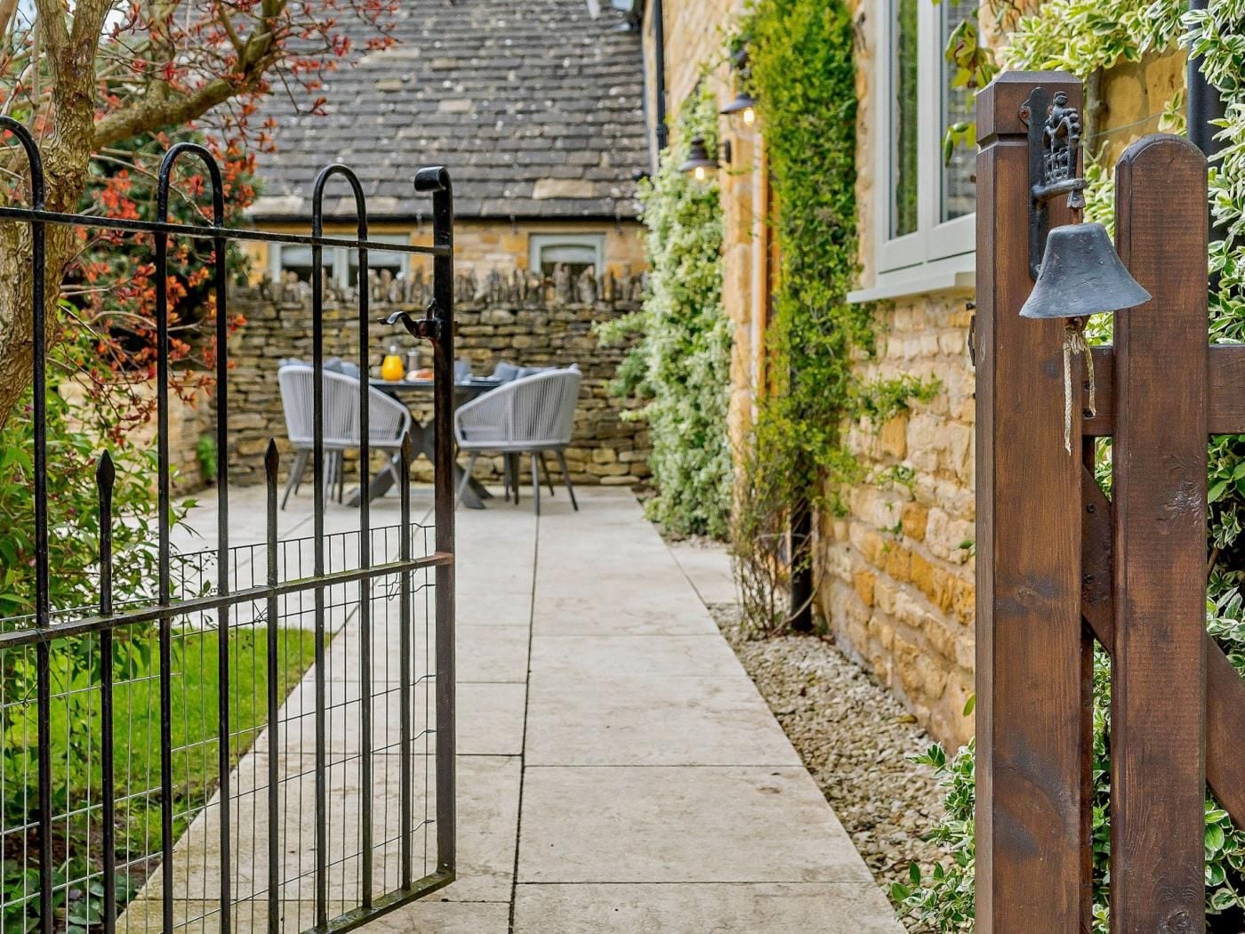A gated entrance to a property with a stone wall, a wooden gate, and a paved pathway leading to a seating area with chairs and a table.