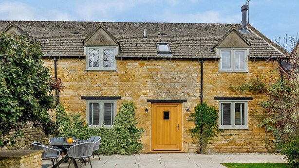 Une maison en pierre avec une porte en bois, deux fenêtres avec volets, une cheminée et une terrasse aménagée avec du mobilier d’extérieur.