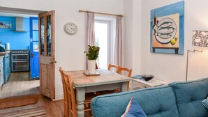 Ein gemütliches Wohnzimmer mit einem blauen Sofa, einem Esstisch mit Stühlen, einer Küche im Hintergrund und einer Uhr an der Wand.
