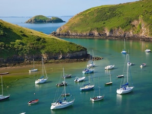A coastal area with multiple sailboats anchored in calm waters, surrounded by green hills and rocky outcrops.