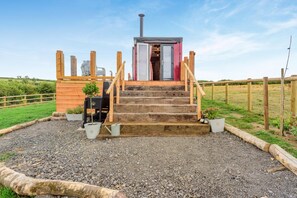 Een houten trap leidt naar een klein gebouw met schoorsteen, omgeven door een grindpad en een houten omheining.