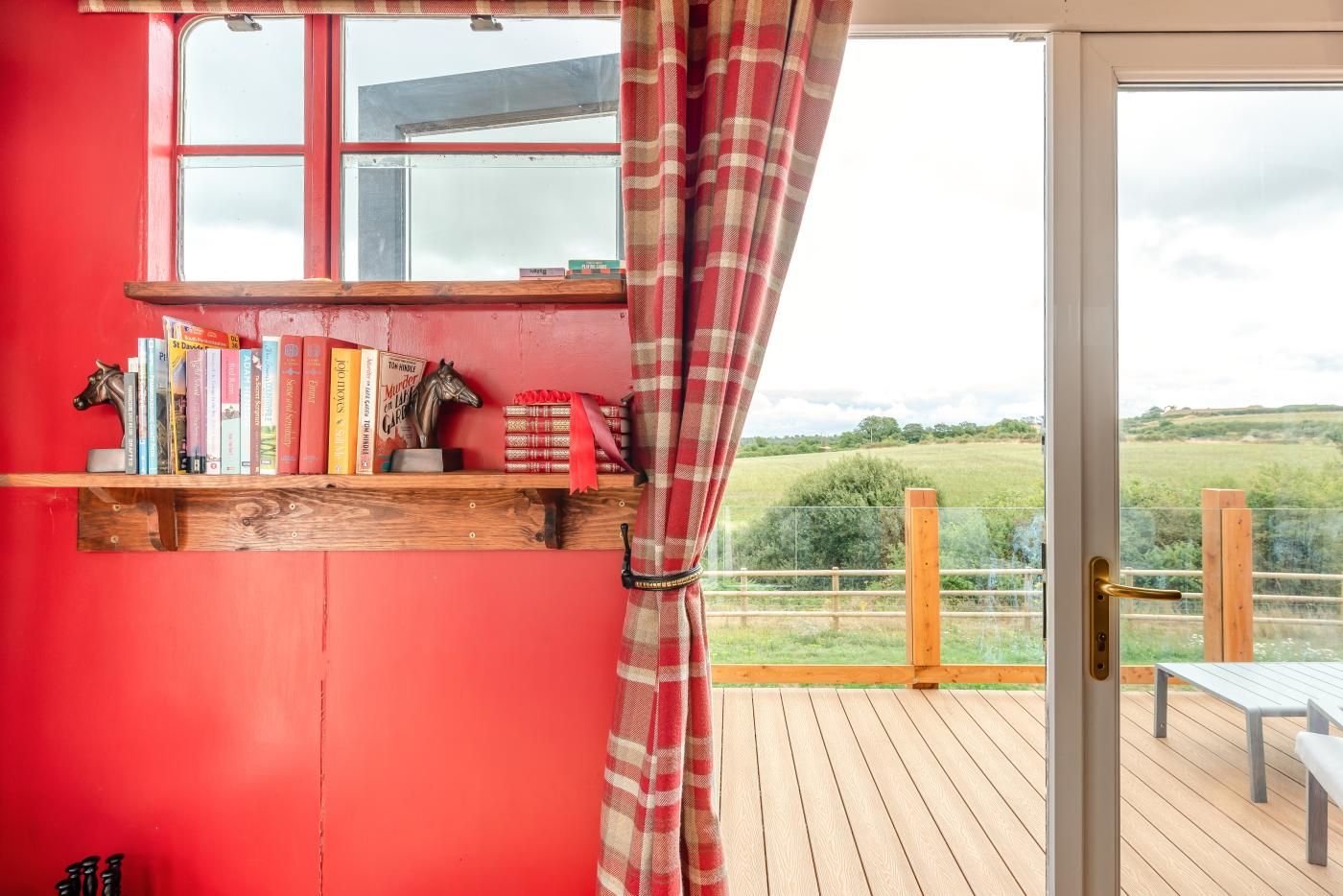 A room with a red wall, a wooden bookshelf, a window with a view of a green landscape, and a door leading outside.