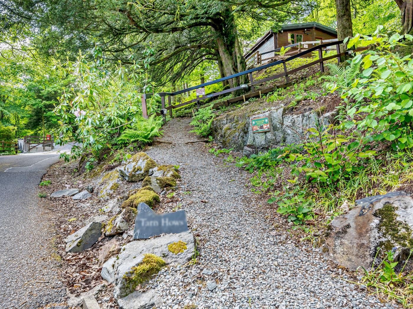 Un chemin de gravier mène à une structure en bois dotée d’une rambarde métallique, le tout étant entouré d’une végétation luxuriante et de rochers.