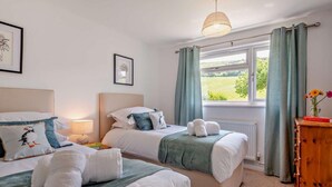 A bedroom with two beds, a bedside table with a lamp, a window with curtains, and a wooden cabinet. - 3 Bed in Wareham (oc-92328) (West Lulworth)
