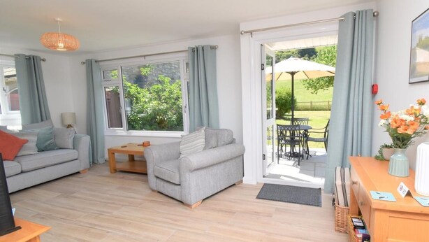 A living room with a sofa, a table, and a view of the garden through a glass door. - 3 Bed in Wareham (oc-92328) (West Lulworth)