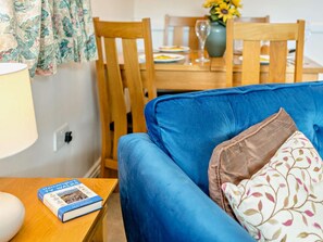 Ein Zimmer mit einem blauen Sofa, einem Holzesstisch mit Stühlen, einer Vase mit Blumen und einem Buch auf einem Beistelltisch.