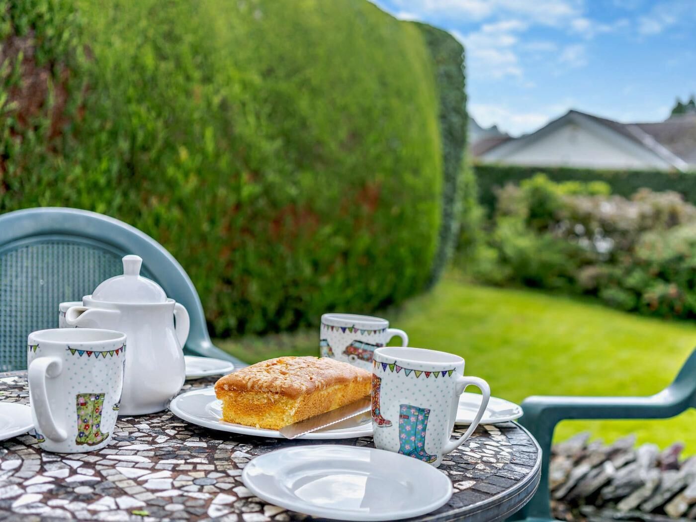 Ein Tisch mit Teekanne, Tassen, einem Teegebäck und einem Teller, draußen aufgestellt, mit einer Hecke und einem Haus im Hintergrund.