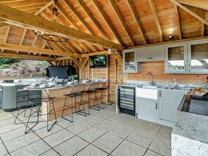 Una cocina exterior con suelo de baldosas de piedra, vigas de madera y una parrilla empotrada.