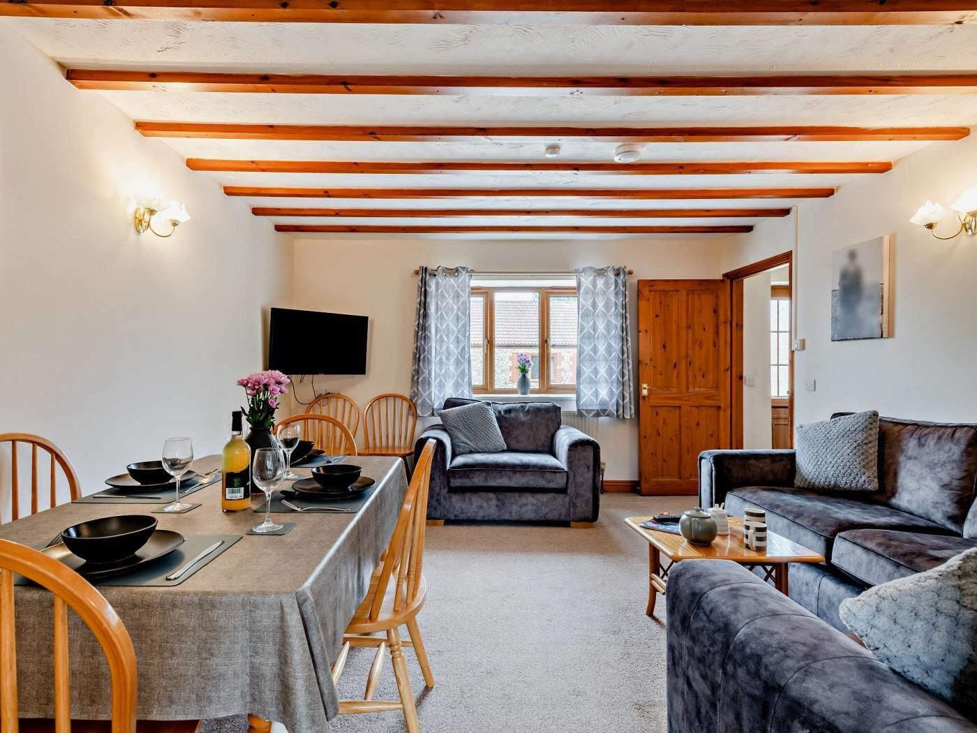 A cozy living room with a dining area, wooden beams on the ceiling, a flat-screen TV, and a window with curtains.