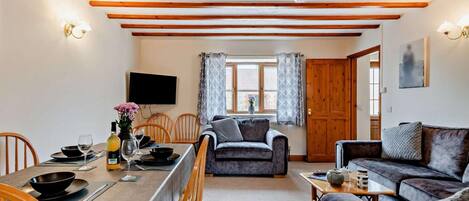 A cozy living room with a dining area, wooden beams on the ceiling, a flat-screen TV, and a window with curtains.