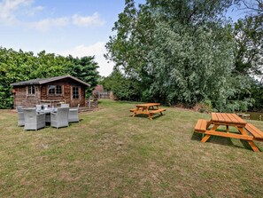 A wooden cabin with a patio area, surrounded by trees and a grassy lawn. - 2 Bed in Dereham (oc-92392) (Horningtoft)