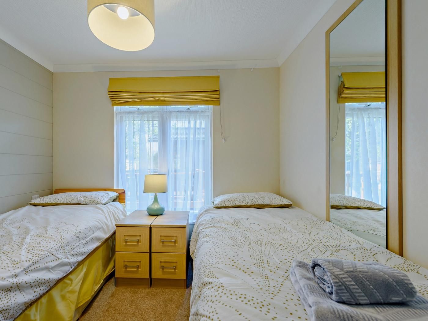 Habitación doble con dos camas, una mesita de noche con lámpara, un espejo y una ventana con cortinas.