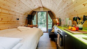 A cozy cabin interior with a bed, kitchenette, and a large window offering a view of the outdoors.