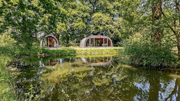 Two modern, semi-open structures with glass walls, surrounded by lush greenery and a calm water body.