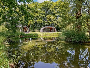 Two modern, semi-open structures with glass walls, surrounded by lush greenery and a calm water body.