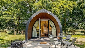 A modern, arched wooden structure with a sign reading 'Valley Pods.' Inside, there is a dining area with a table and chairs.