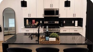 Une cuisine moderne avec des meubles blancs, un plan de travail noir, un four à micro-ondes en acier inoxydable et un coin dédié au café et au thé.