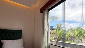 A hotel room with a bed, a window with a view of greenery and a tower, and an air conditioning unit.