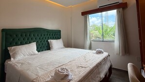 A hotel room with a large bed, a green headboard, a window with curtains, an air conditioning unit, and a small table with a chair.