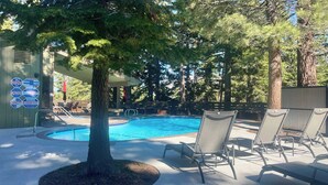 A swimming pool with lounge chairs, a tree, and a building with safety signs. - Cozy Village Condo | Hot Tub, Sauna, Fireplace (Mammoth Lakes)