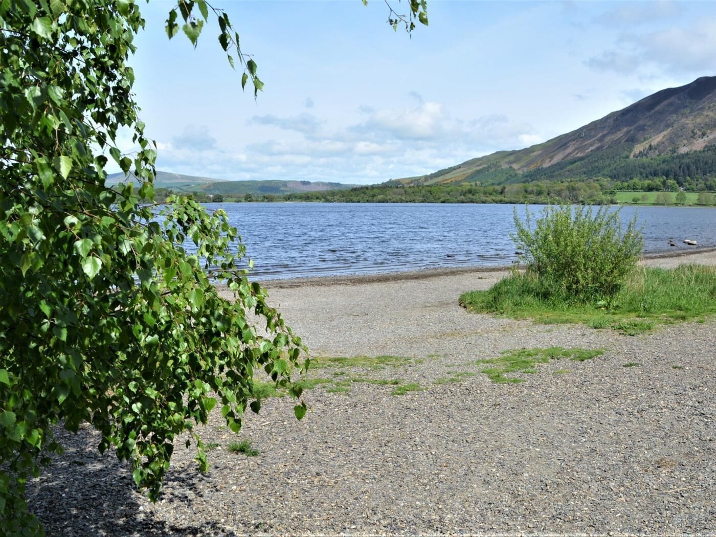 Een kiezelstrand met een meer, omringd door heuvels en begroeiing.