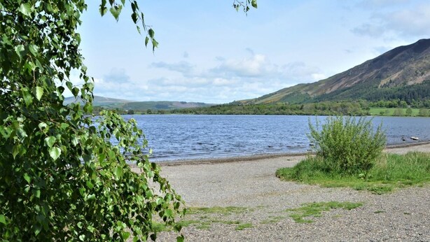 Ein Kiesstrand mit einem See, umgeben von Hügeln und viel Grün.