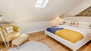 Un dormitorio con techo inclinado, una cama con colcha amarilla y azul, una pequeña silla con cojín, una mesita de noche con lámpara y un tragaluz.