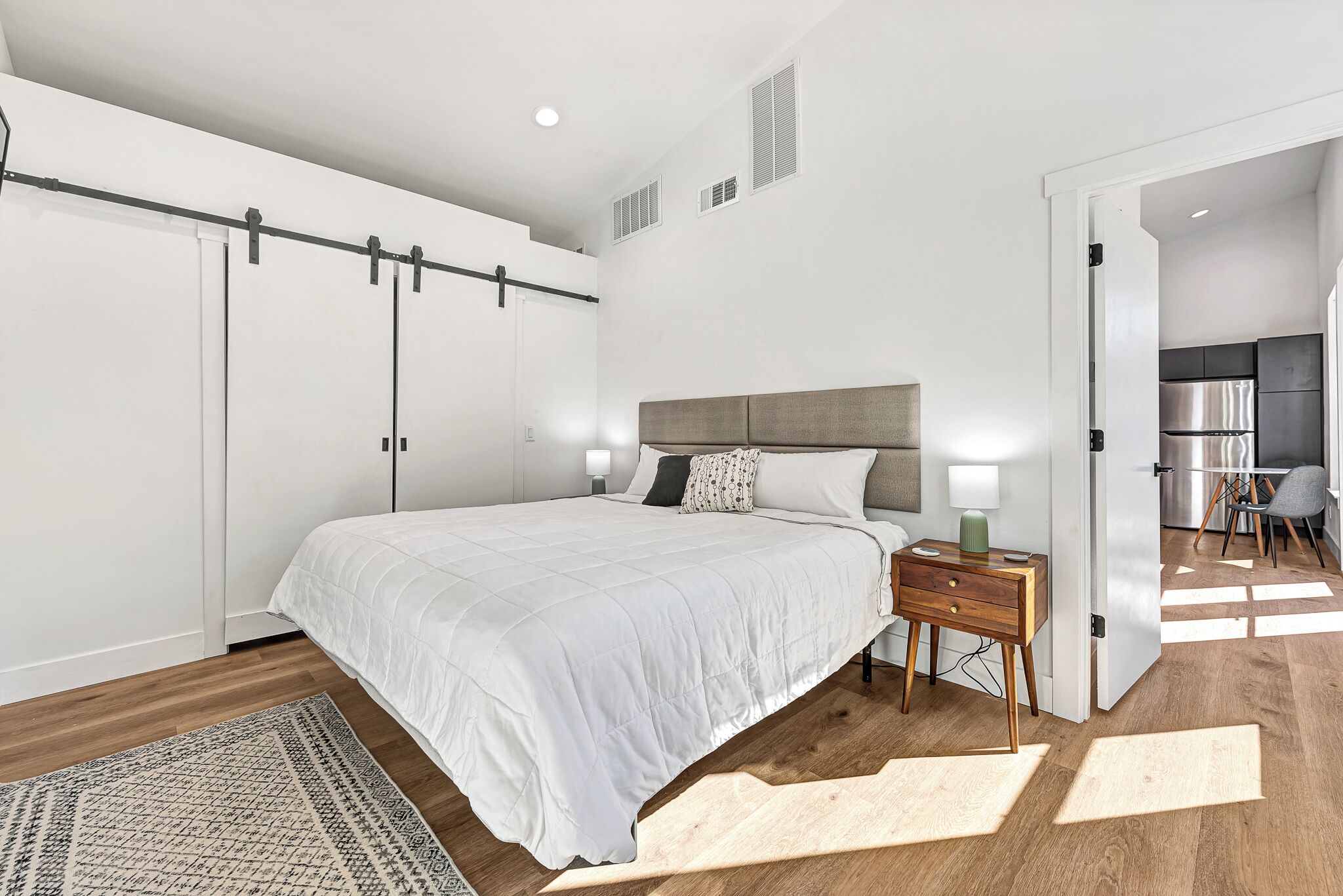 Un dormitorio moderno con una cama grande, una mesita de noche, piso de madera y vista a la cocina.