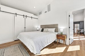 Ein modernes Schlafzimmer mit einem großen Bett, einem Nachttisch, Holzboden und Blick in die Küche.