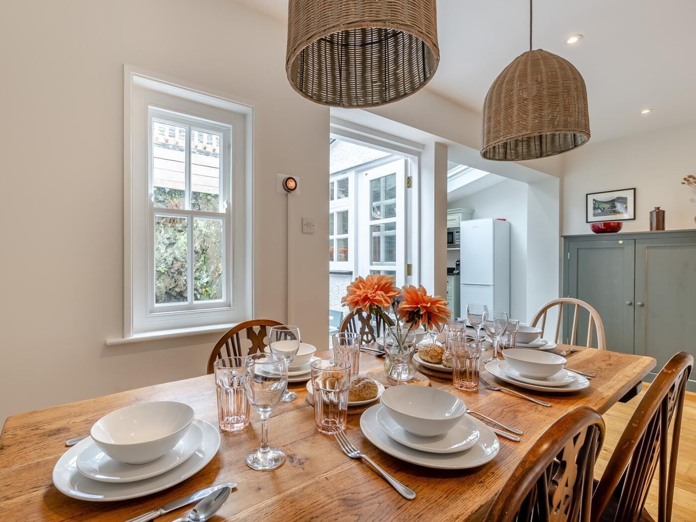 A dining area with a wooden table set for four, surrounded by chairs, with hanging wicker lamps and a view of the outdoors through a window.