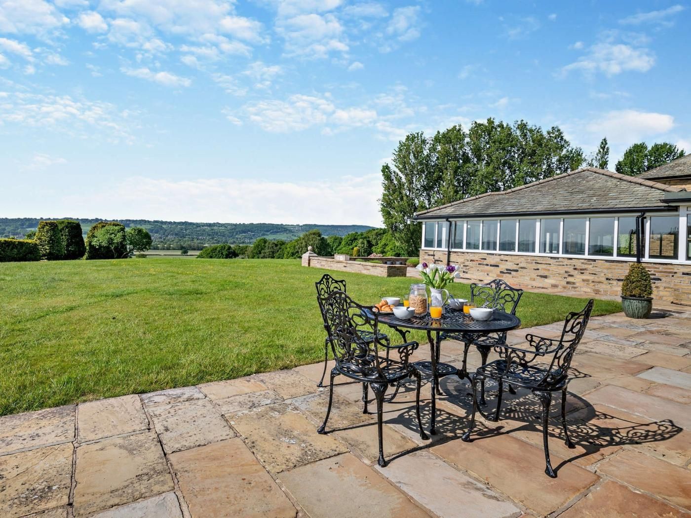 Un patio al aire libre con una mesa dispuesta para el desayuno, rodeado de suelo de piedra y vistas a un campo cubierto de césped y una casa con invernadero.