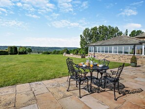 An outdoor patio with a table set for breakfast, surrounded by stone flooring, and overlooking a grassy field and a house with a conservatory. - 2 Bed in Otley (oc-92378) (Leathley)