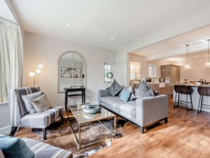 Ein modernes Wohnzimmer mit Sofa, Ohrensessel und Couchtisch, im Hintergrund eine Küche und ein Essbereich mit Barhocker.