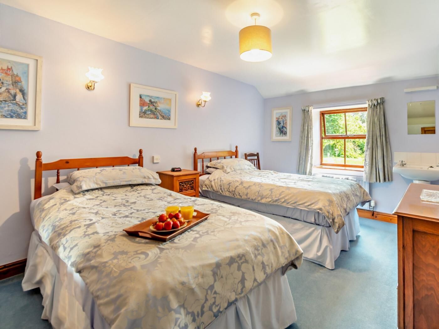 A bedroom with two beds, a bedside table with a tray of fruit, a window with curtains, and a bathroom with a sink and mirror.