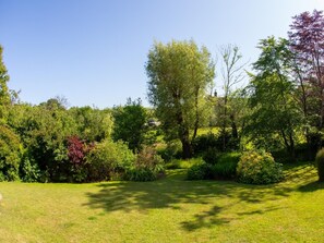 Ein gepflegter Garten mit verschiedenen Bäumen und Sträuchern, einem deutlichen Weg und einem Gebäude im Hintergrund.