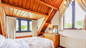 Een slaapkamer met een schuine plafond, een bed met een witte spreideken, een houten dressoir en een raam met gordijnen.