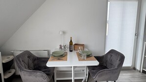 Un coin repas avec une table blanche, deux chaises grises, une bouteille, des verres et un panier.