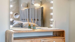 A vanity with a mirror, lights, and a bed in the background.
