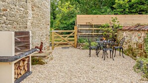 Een stenen gebouw met een barbecue, een houten poort en een tafel met stoelen in een grindpad.